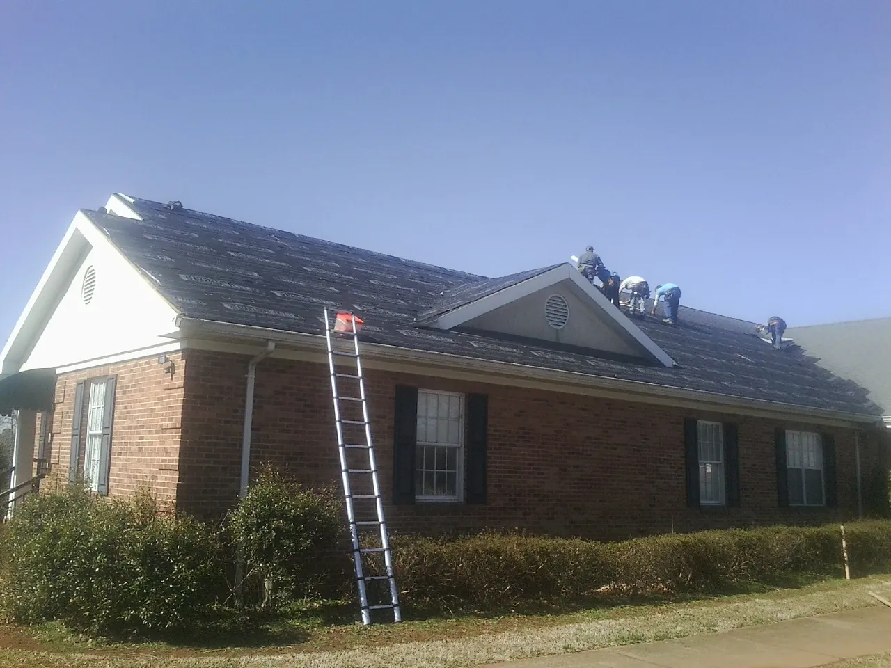 Crew tearing shingles off a brick ranch roof with a ladder set — Emergency Roof Repair work by our crew in Four Lakes