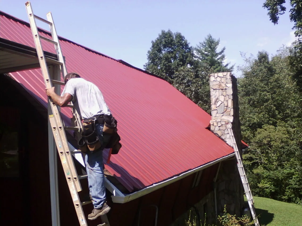 Roofer on a ladder beside a red metal roof and stone chimney — Soffit & Fascia Repair by Roofing Brothers in Four Lakes